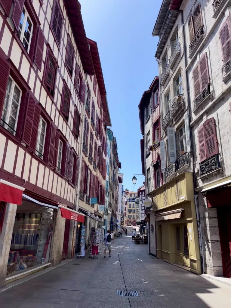 Blick durch eine enge Gasse in der Altstadt von Bayonne mit markanten, traditionellen Fachwerkhäusern mit weinroten Holzelementen und Fensterläden unter strahlend blauem Himmel.