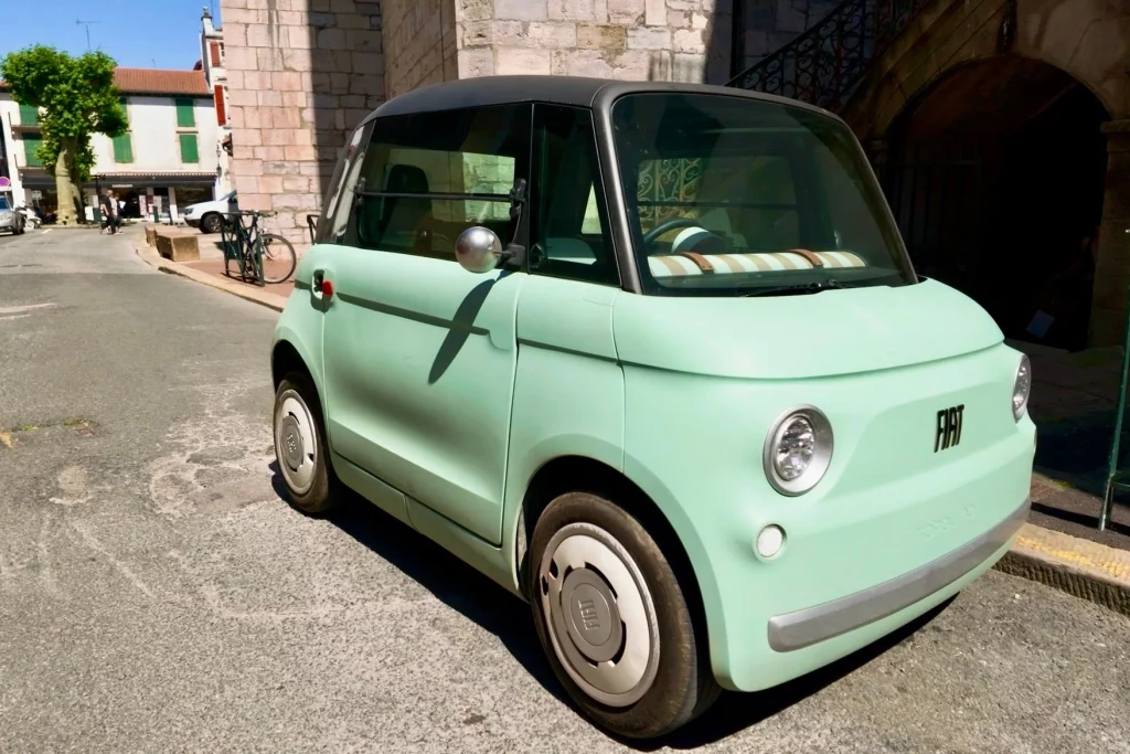 Ein kleiner, moderner Fiat Topolino in Mintgrün steht geparkt in einer historischen Gasse der Altstadt von Bayonne vor einer alten Steinmauer.