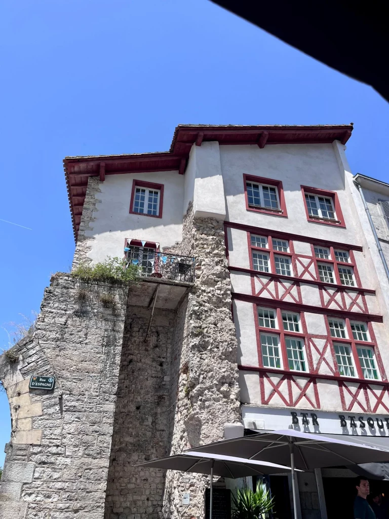 Blick aus einem Straßencafé auf das markante Gebäude der "Bar Basque" in der Rue d'Espagne in Bayonne, dessen weiße Fassade mit rotem Fachwerk direkt an Überreste einer historischen, hohen Steinmauer und einen kleinen Balkon grenzt.