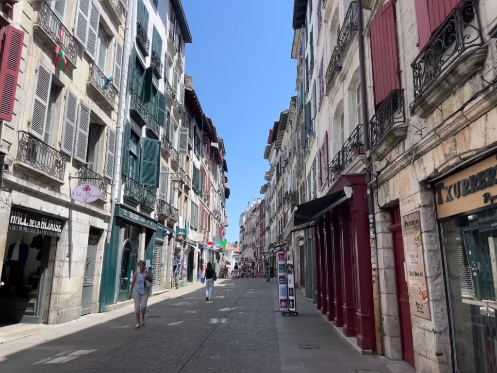 Eine gemütliche, gepflasterte Einkaufsstraße in Bayonne unter blauem Himmel, gesäumt von historischen mehrstöckigen Gebäuden mit farbenfrohen Fensterläden in Grün und Weinrot.