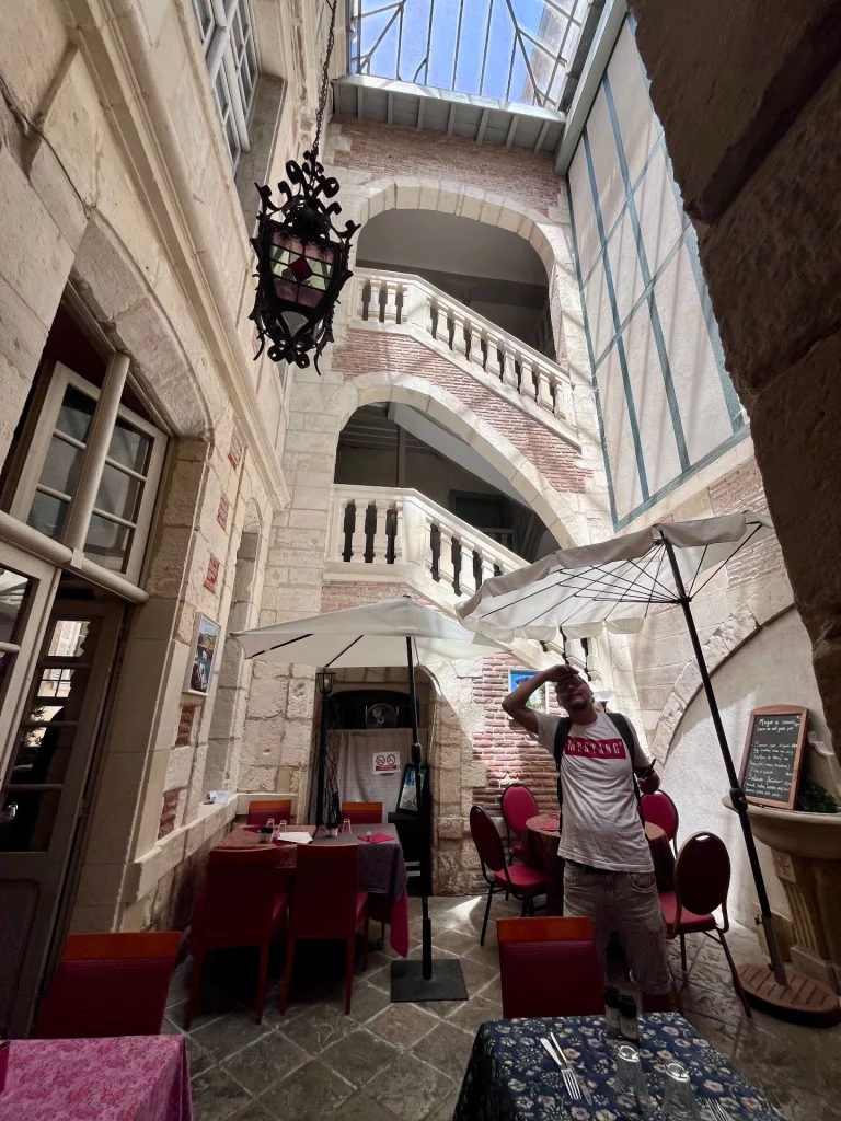 Ein beeindruckender, mehrstöckiger Innenhof eines historischen Gebäudes in Bayonne mit geschwungenen Steintreppen, einer großen Hängelaterne und einem gemütlichen Restaurantbereich mit Sonnenschirmen im Erdgeschoss.