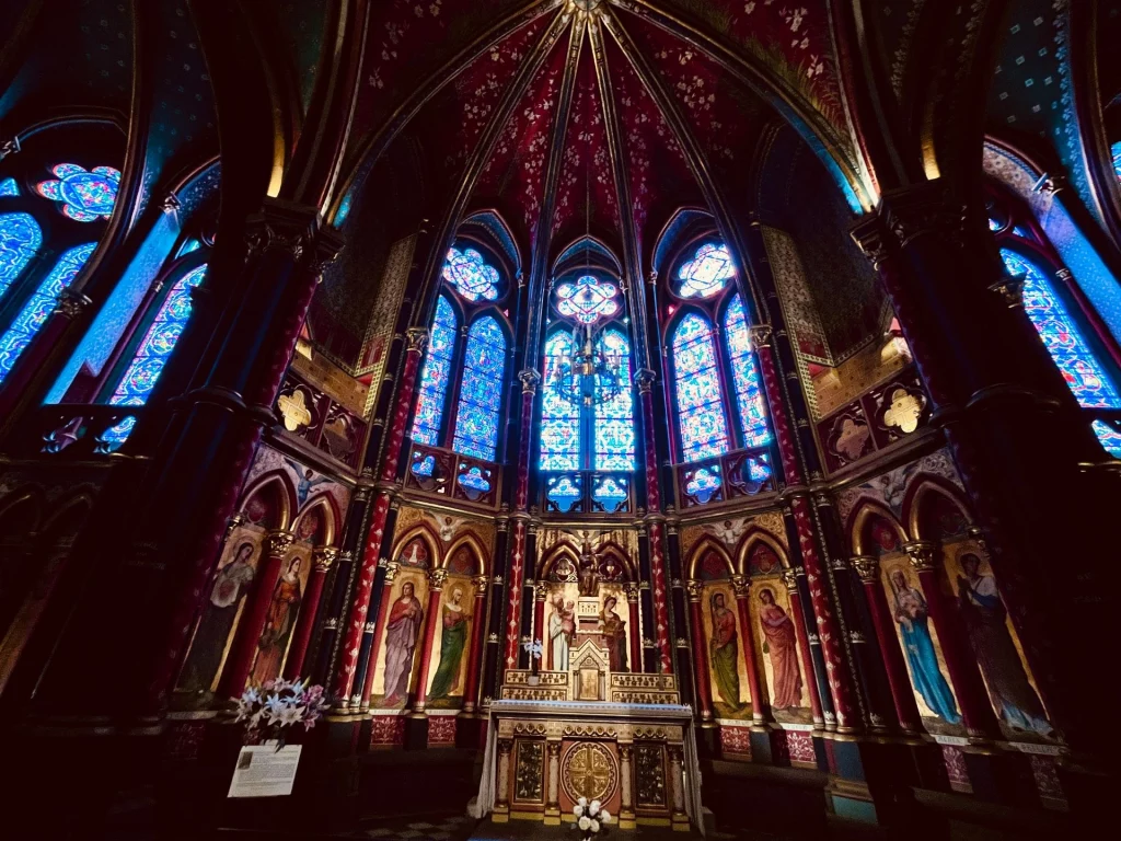 Der prachtvolle, in Dunkelrot und Gold gestaltete Altarraum der Kathedrale von Bayonne unter einem dramatischen, gotischen Deckengewölbe und leuchtend blauen Kirchenfenstern.