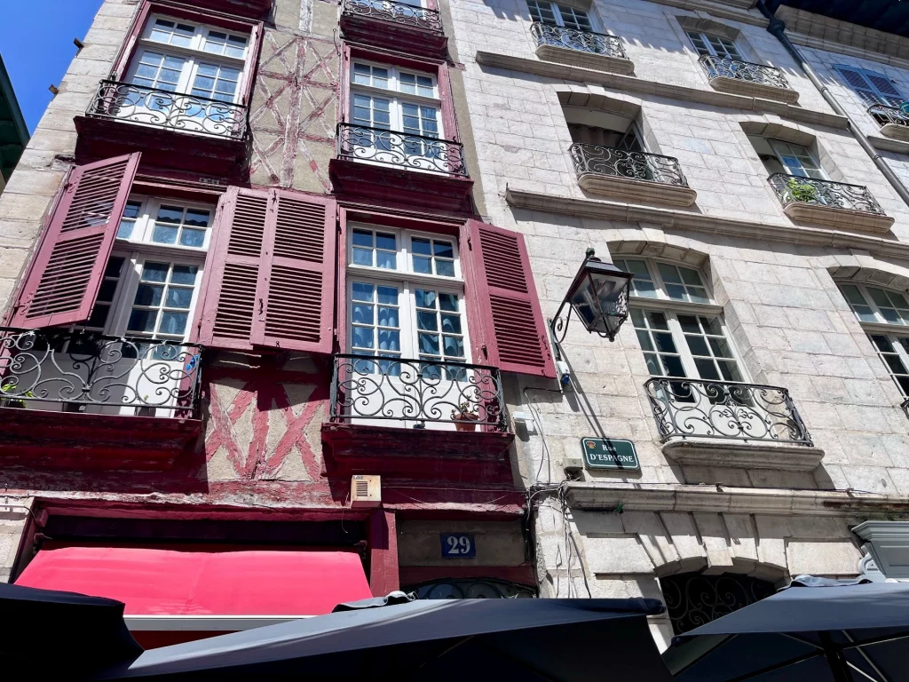 Detailaufnahme einer historischen Hausfassade in der Rue d'Espagne in Bayonne mit weinroten Fensterläden, gusseisernen Balkongittern und einer klassischen Straßenlaterne.