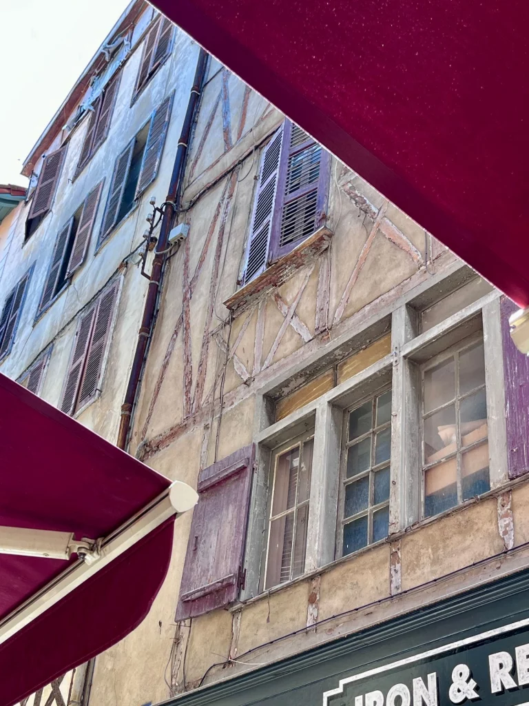 Blick nach oben aus einem Straßencafé in Bayonne, teilweise eingerahmt von einer weinroten Markise, auf eine charmante, wettergegerbte historische Fachwerkfassade mit hölzernen Fensterläden.