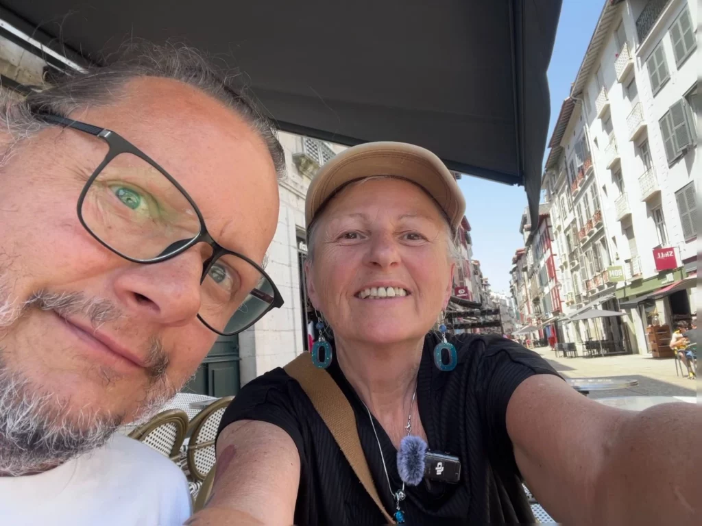 Ein fröhliches Selfie von Ingo mit seiner Brille und der lächelnden Karin mit beiger Kappe, die unter einem Sonnenschirm in einem Straßencafé in Bayonne sitzen; im Hintergrund ist eine helle Häuserzeile der Altstadt zu sehen.