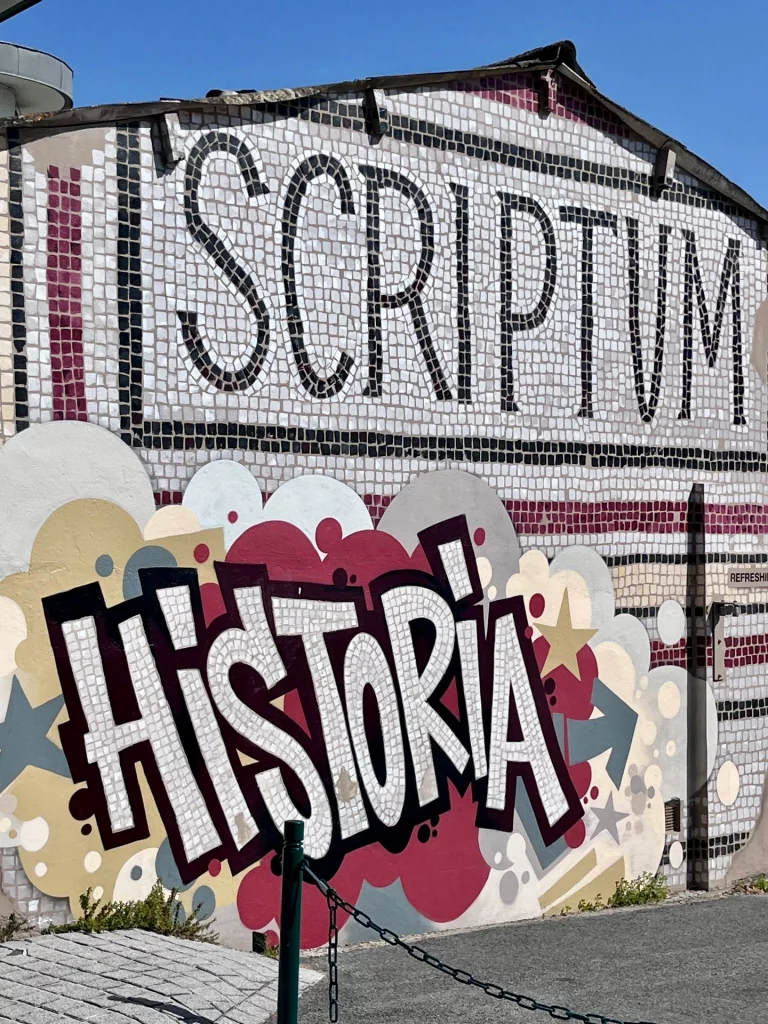 Eine kunstvoll gestaltete Hauswand in Bayonne mit einem großen Mosaik-Schriftzug "SCRIPTUM" und einem modernen Graffiti-Schriftzug "HISTORIA" im Streetart-Stil.