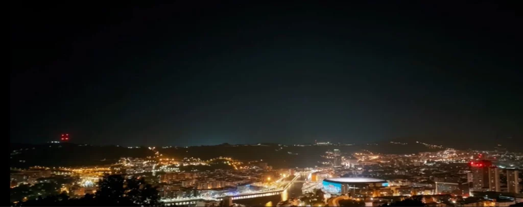 Weites Nachtpanorama über die beleuchtete Stadt Bilbao, das San-Mamés-Stadion mit blauem Lichtring und die umliegenden dunklen Hügel im Hintergrund.