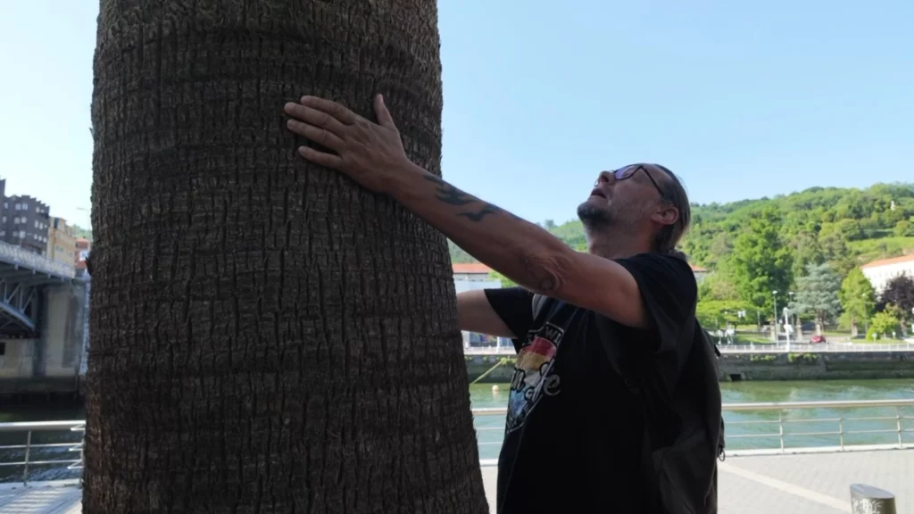 Ingo mit Brille und Tätowierung am Arm steht am Flussufer und lehnt sich mit ausgestreckten Armen an den Stamm einer großen Palme.