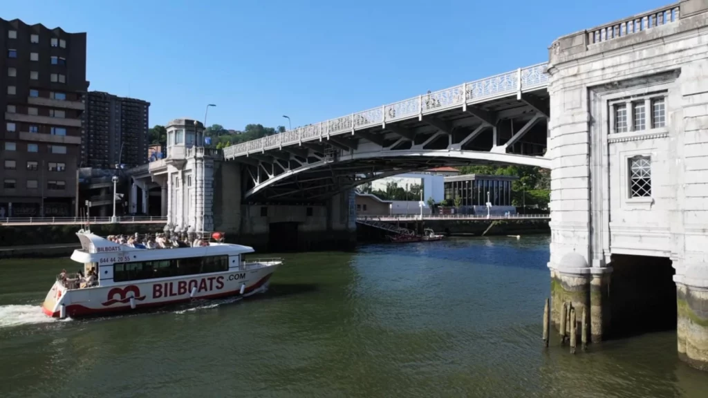 Ein Ausflugsboot mit der Aufschrift „BILBOATS“ fährt auf dem Fluss Nervión unter einer großen Bogenbrücke hindurch.