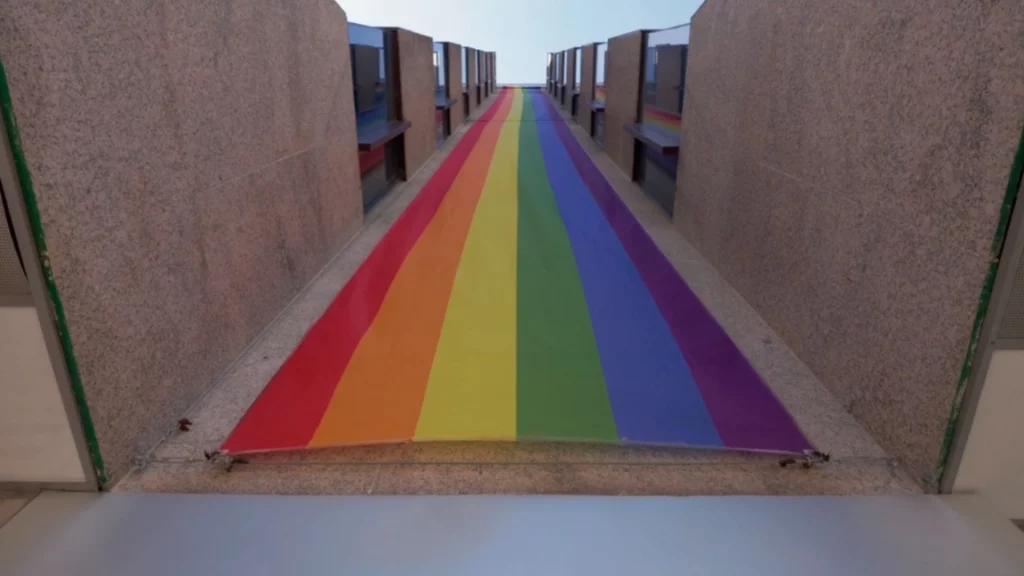 Eine lange Regenbogenfahne ist flach auf einer Rampe oder einem Durchgang zwischen zwei modernen Betonwänden ausgebreitet.