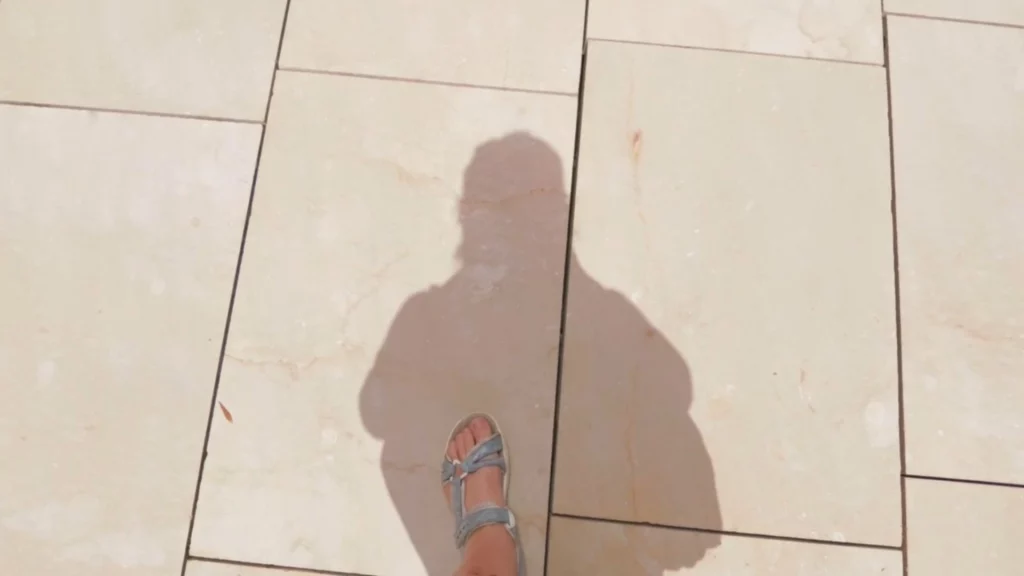 Blick von oben auf helle Steinbodenfliesen, auf denen der Schatten einer gehenden Person mit Sandalen zu sehen ist.