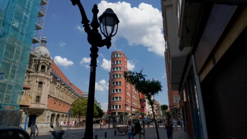 Blick durch eine Straße im Stadtzentrum von Bilbao auf ein markantes, rötliches, schmales Gebäude an einer Kreuzung, im Vordergrund eine historische Straßenlaterne.