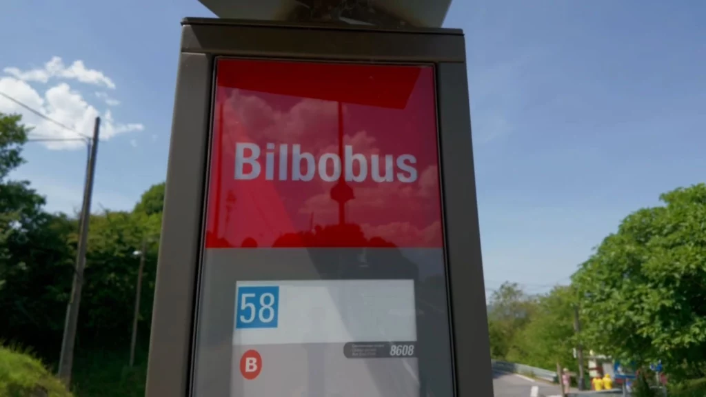 Nahaufnahme eines roten Schildes mit der weißen Aufschrift "Bilbobus" an einer Bushaltestelle vor blauem Himmel und Bäumen.