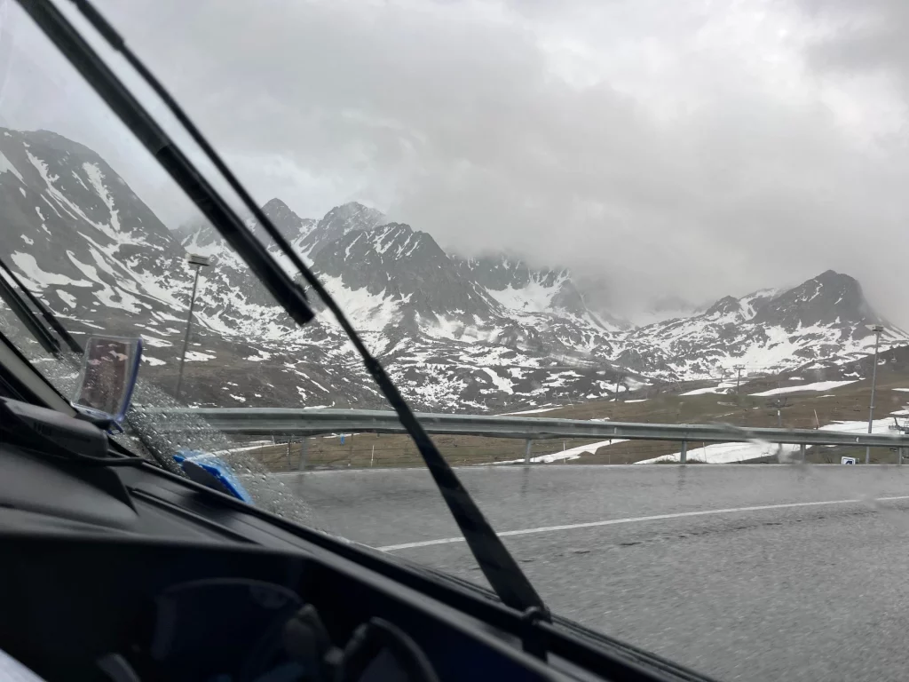 Blick aus der Windschutzscheibe eines Wohnmobils mit Scheibenwischern in Aktion auf eine nasse Straße und schneebedeckte Berghänge im Nebel.