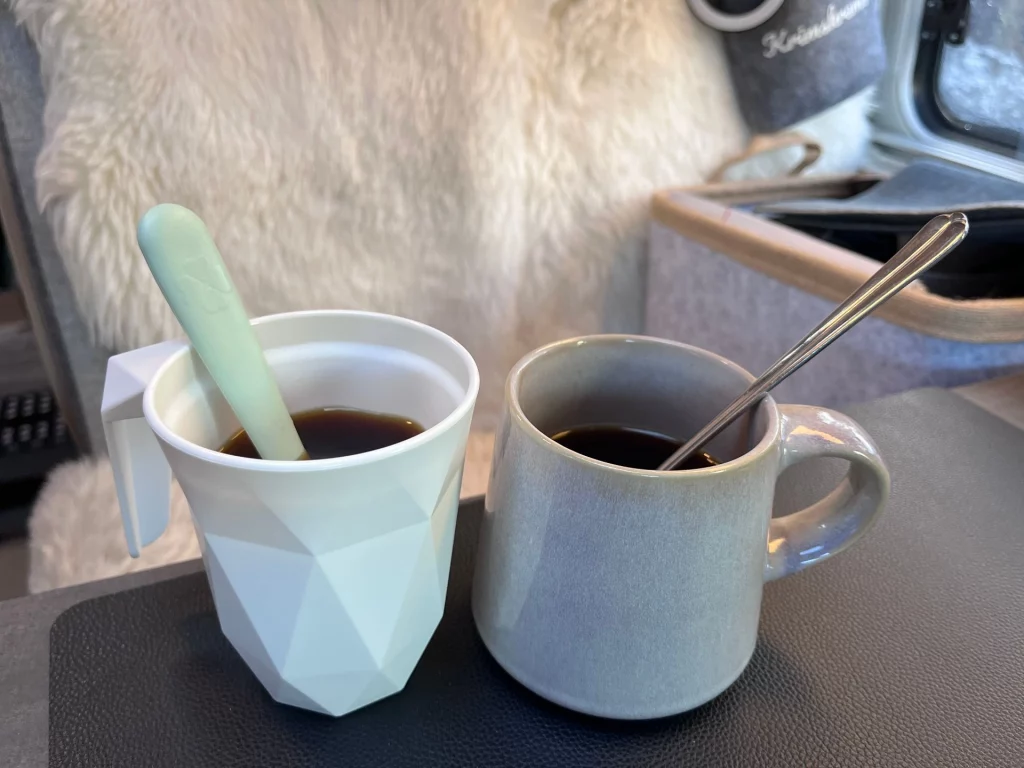 Zwei gefüllte Kaffeetassen mit Löffeln stehen auf dem Tisch im gemütlich eingerichteten Innenraum des Wohnmobils vor einem hellen Lammfell.