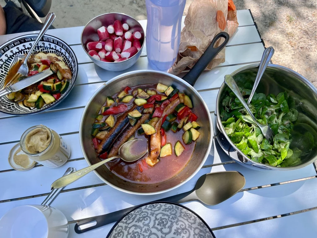 Ein Campingtisch mit einer Pfanne gebratenem Gemüse und veganen Würstchen, einer Schüssel frischem Ackersalat, Radieschen und Senf.
