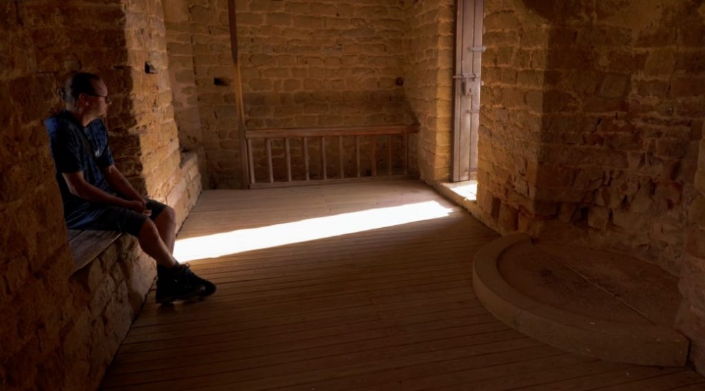 Ingo sitzt im tiefen Schatten auf einer Steinbank in einem historischen Raum des Castillo de Loarre, während ein heller Sonnenstrahl über den Holzboden wandert.
