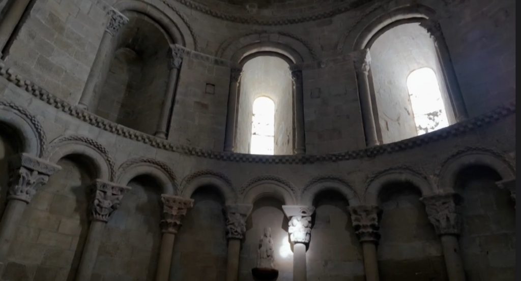 Das schattige Innere der romanischen Kirche San Pedro im Castillo de Loarre mit Rundbögen, verzierten Kapitellen und Tageslicht, das durch die hohen Rundbogenfenster fällt.
