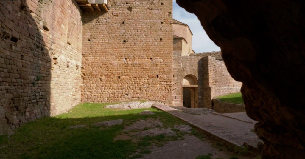 Blick aus dem Schatten eines Felsens auf die hohen, sonnenbeschienenen Sandsteinmauern und einen inneren Torbogen der Burg Loarre.