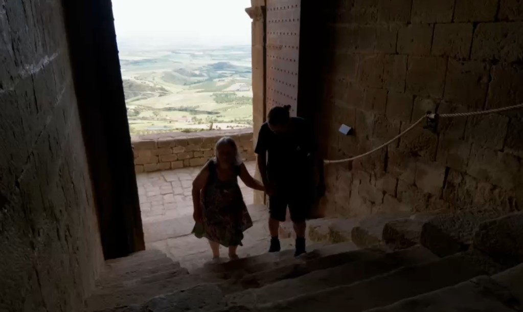 Blick von einer dunklen Steintreppe im Castillo de Loarre nach draußen durch ein großes, geöffnetes Burgtor auf die weite, helle Landschaft.