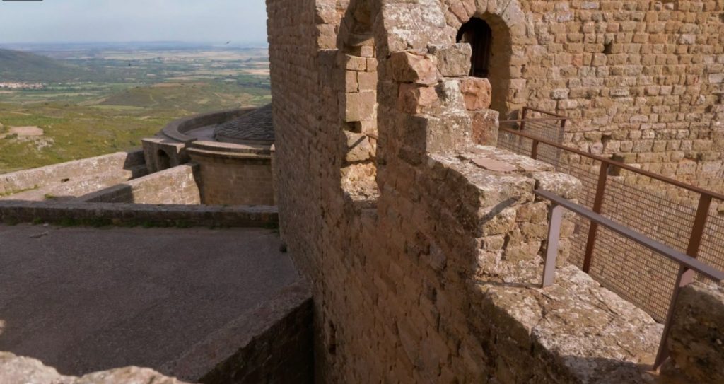 Blick von oben entlang einer steinernen Wehrmauer mit Rundbogenfenstern im Castillo de Loarre auf die tiefer gelegenen Burgteile und das grüne Tal im Hintergrund.