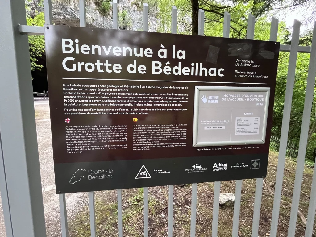Ein braunes Informationsschild mit der Aufschrift Bienvenue à la Grotte de Bédeilhac an einem grauen Metallgitterzaun vor dem Höhleneingang.