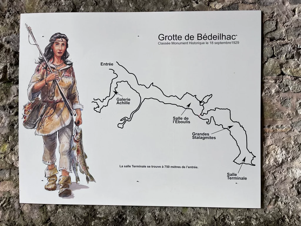 Eine weiße Infotafel an einer Steinwand zeigt den gezeichneten Verlauf der Grotte de Bédeilhac und eine Illustration einer eiszeitlichen Jägerin mit Fischen.