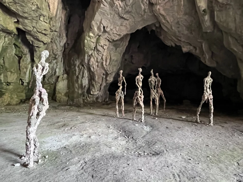 Mehrere abstrakte, menschengroße Skulpturen aus lehmartigem Material stehen im sandigen Boden einer großen, hellen Felshöhle.