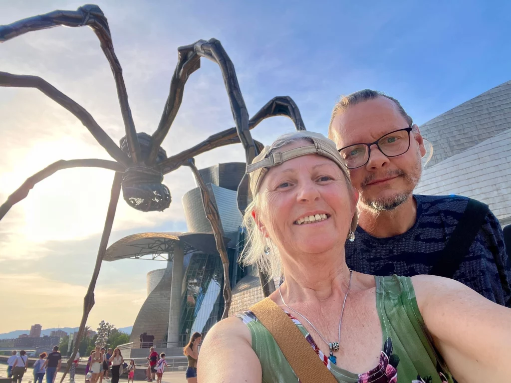 Ein lächelndes Paar macht ein Selfie vor der riesigen Spinnenskulptur Maman am Guggenheim Museum in Bilbao bei tiefstehender Sonne.
