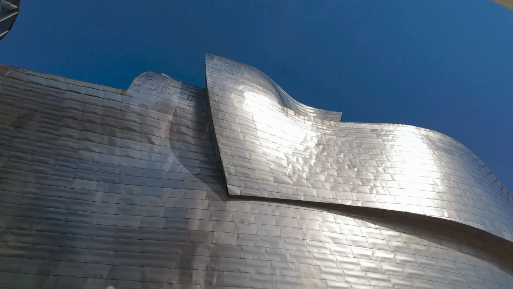 Nahaufnahme der wellenförmigen, silbernen Titanplatten des Guggenheim-Museums in Bilbao, die das Sonnenlicht reflektieren, vor blauem Himmel.