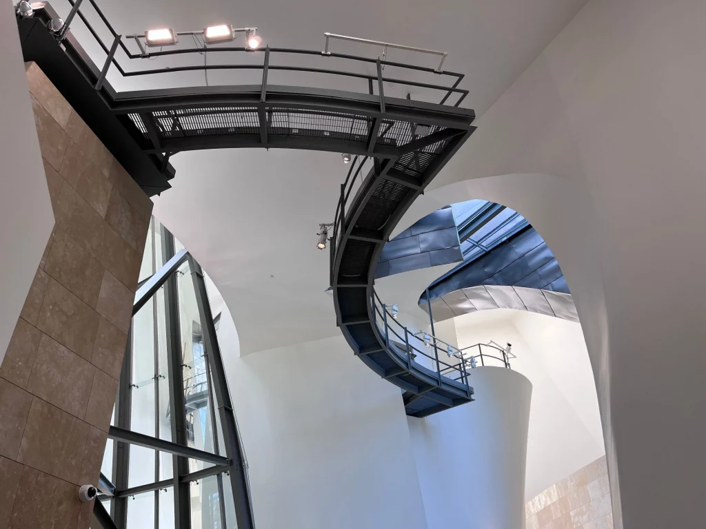 Blick nach oben in das helle Atrium des Guggenheim-Museums mit geschwungenen weißen Wänden und einer frei schwebenden, dunklen Metall-Laufbrücke.