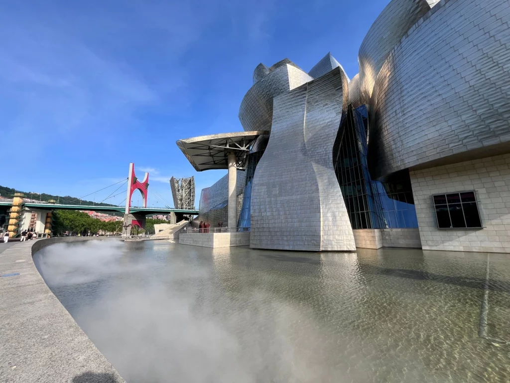 Das Guggenheim-Museum Bilbao mit seinen geschwungenen Titanplatten im Tageslicht, im Vordergrund künstlicher Nebel auf einer Wasserfläche und die rote La-Salve-Brücke im Hintergrund.