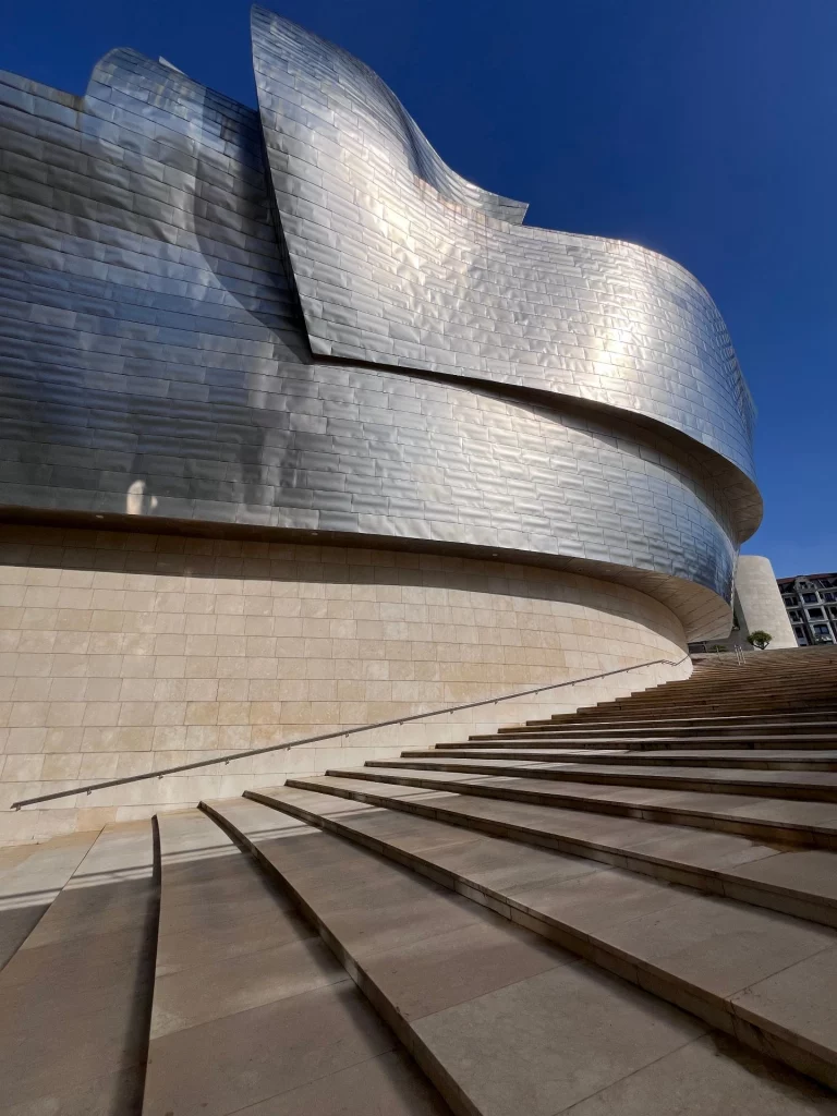 Blick von unten auf die geschwungene, glänzende Titanfassade des Guggenheim-Museums in Bilbao mit einer breiten Steintreppe im Vordergrund unter blauem Himmel.