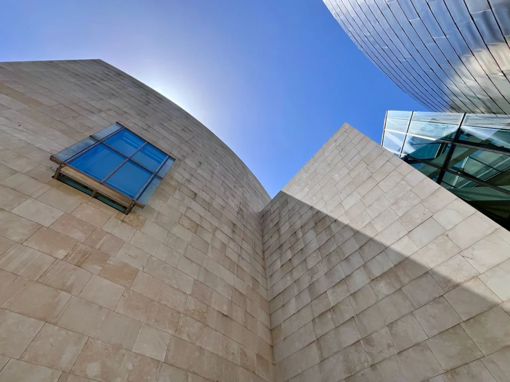 Eine kreative Architekturfotografie aus der Froschperspektive, die die hellen Kalksteinwände, ein blau spiegelndes Fenster und die Titanplatten des Guggenheim-Museums vor blauem Himmel zeigt.
