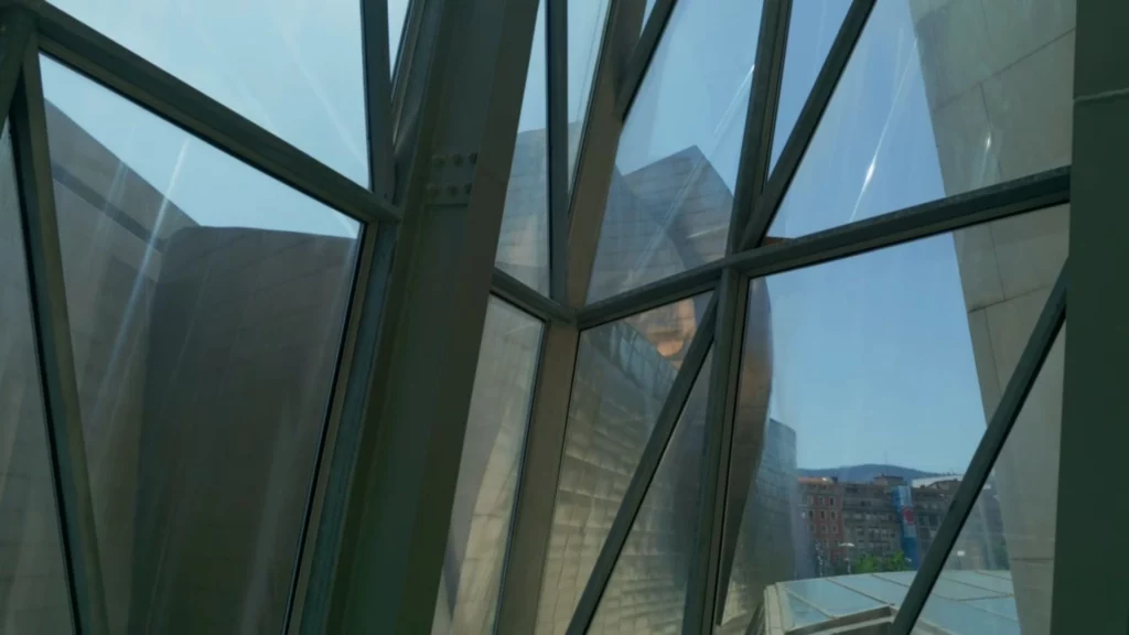 Blick durch die geometrischen Metallverstrebungen einer riesigen Glasfassade im Guggenheim-Museum auf die gegenüberliegenden Titanplatten und die Stadt im Hintergrund.