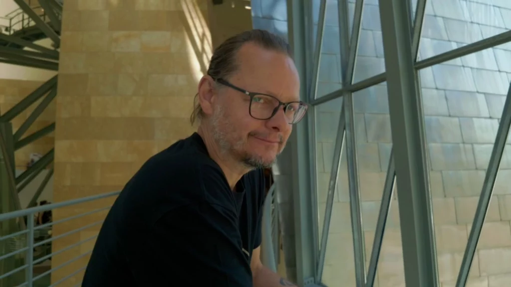 Porträt von Ingo mit Brille, der sich im Inneren des Museums an eine riesige Glasfassade lehnt und lächelnd in die Kamera blickt.