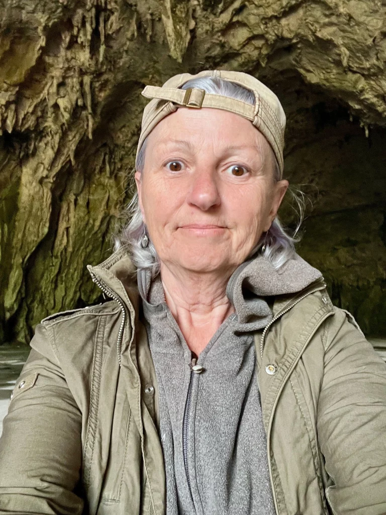 Selfie von Karin Schmidtke mit einer khakifarbenen Jacke, Fleece-Pulli und einer umgedrehten Basecap vor dem steinernen Hintergrund der Grotte de Bédeilhac.