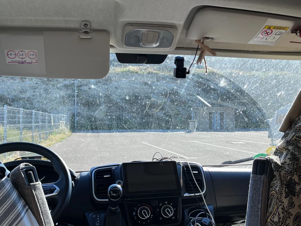 Blick aus dem Cockpit eines Kastenwagens durch die leicht staubige Windschutzscheibe auf den sonnigen Stellplatz.