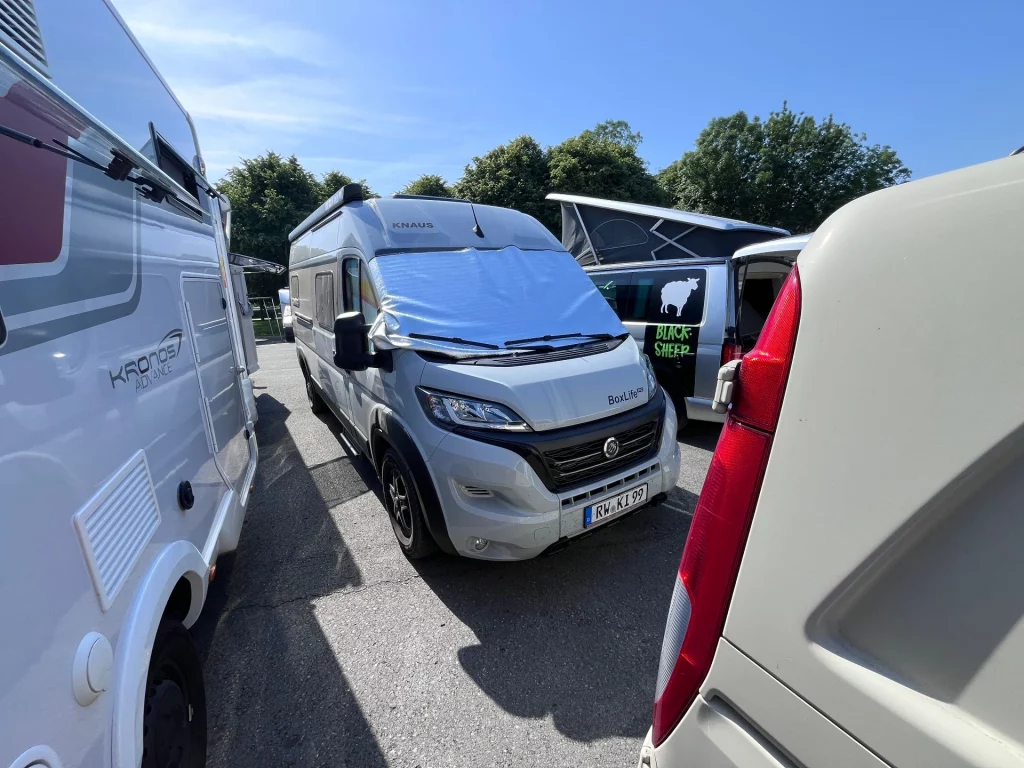 Ein grauer Knaus Box Life Kastenwagen steht mit Thermo-Scheibenmatte auf der Windschutzscheibe auf einem gut besuchten Wohnmobilstellplatz zwischen anderen Campern.