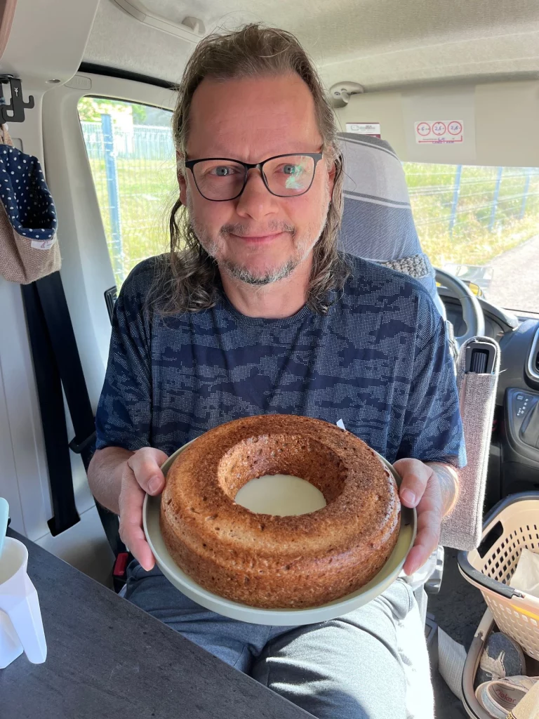 Ingo sitzt lächelnd im Fahrerhaus des Kastenwagens und präsentiert stolz den frisch gebackenen, ringförmigen Kuchen auf einem Teller.