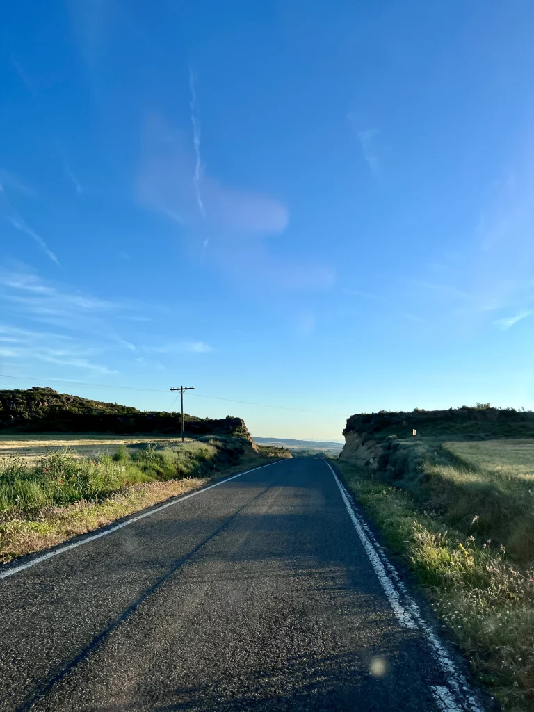 Eine einsame, asphaltierte Landstraße führt bei klarem, blauem Himmel durch eine hügelige Felderlandschaft in Spanien in Richtung Horizont.