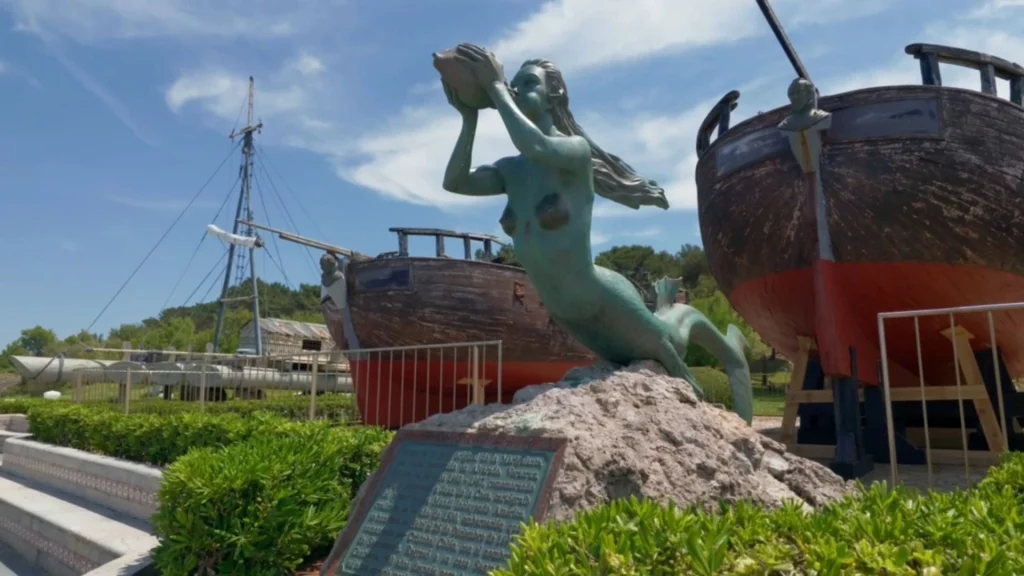 Eine bronzene Meerjungfrau-Statue, die in eine Muschel bläst, im Vordergrund einer Freiluft-Ausstellung mit zwei alten, hölzernen Schiffen aufgebockt an Land unter blauem Himmel.