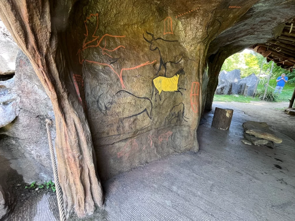 Eine künstliche Felswand im prähistorischen Park mit farbigen Nachbildungen von eiszeitlichen Tierzeichnungen wie Bisons, Pferden und Hirschen in Schwarz und Rot.