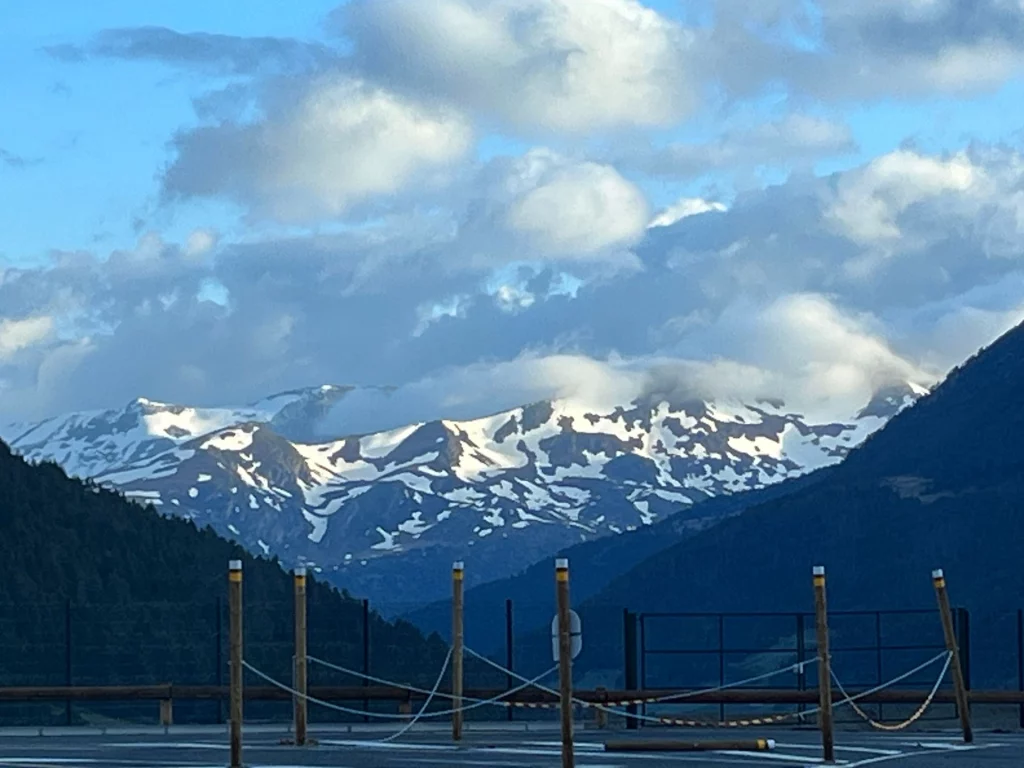 Blick über eine hölzerne Absperrung auf eine mächtige, schneebedeckte Bergkette, deren Gipfel teilweise in dichte Wolken gehüllt sind.