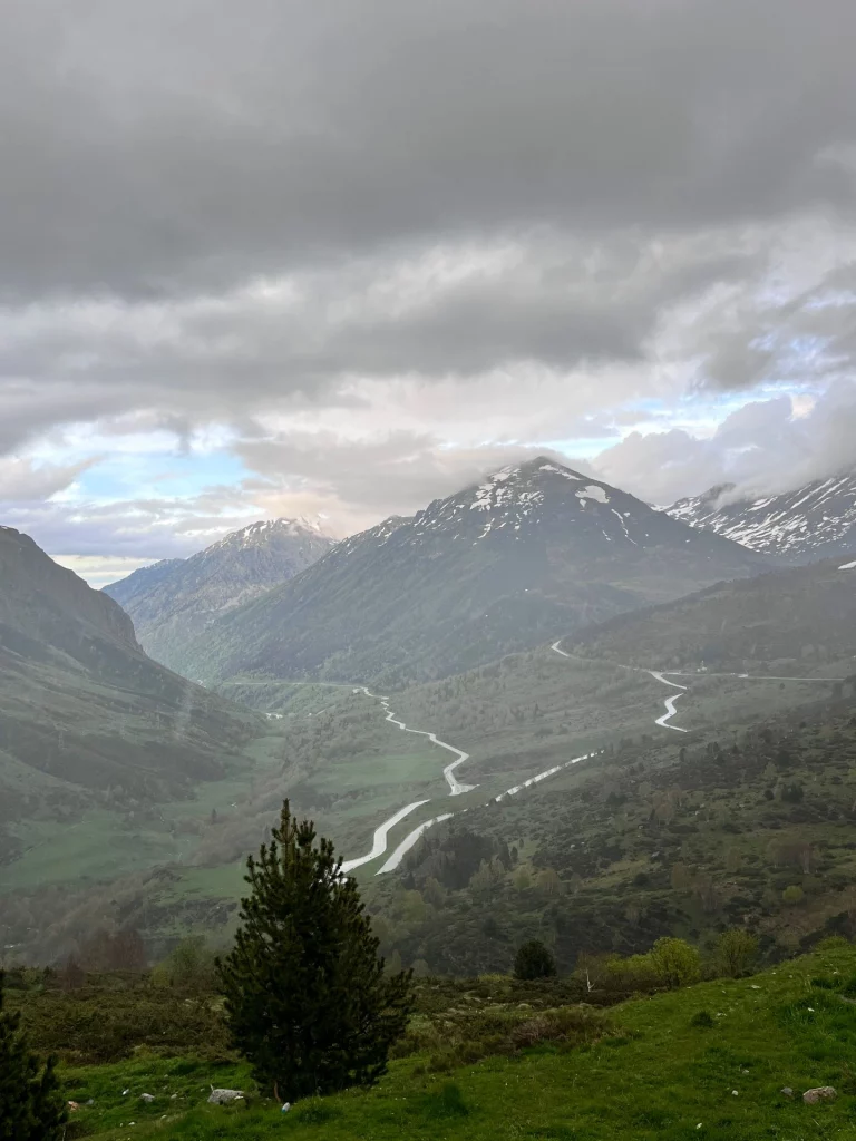 Blick von oben auf eine kurvenreiche Serpentinenstraße, die sich durch ein grünes Tal in den Pyrenäen windet, im Hintergrund schneebedeckte Gipfel.