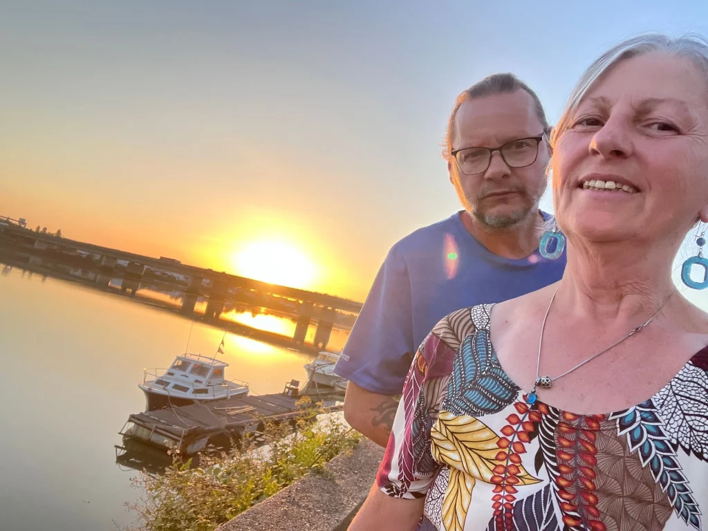 Ingo und Karin stehen lächelnd am Ufer des Flusses Adour in Bayonne während eines malerischen Sonnenuntergangs über dem Wasser.