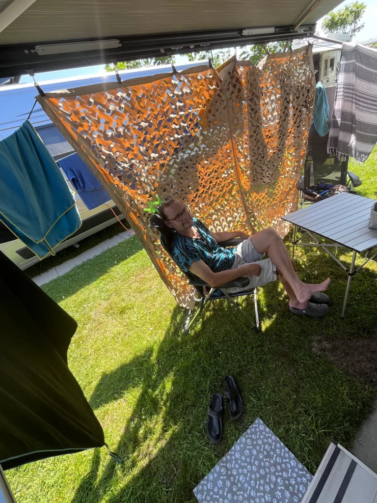 Ein Mann sitzt entspannt im Campingstuhl im Schatten der Markise neben einem Tarnnetz-Sichtschutz.