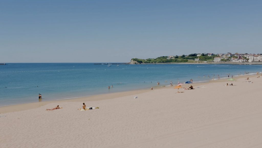 Weite Panoramaaufnahme eines hellen Sandstrandes unter einem klaren, blauen Himmel. Im ruhigen, blauen Meer baden einige Personen, während andere sich auf dem Strand sonnen oder unter bunten Sonnenschirmen entspannen. Im Hintergrund erstreckt sich eine grüne Landzunge, an deren Hang sich eine Ortschaft mit hellen Gebäuden befindet.