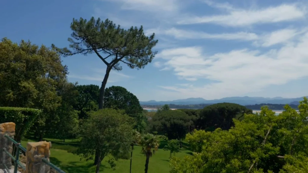 Weiter Ausblick von einer Steinterrasse über eine dichte, grüne Parklandschaft mit Pinien und Palmen. Im Hintergrund erstrecken sich Hügelketten und das Meer unter leicht bewölktem, blauem Himmel.