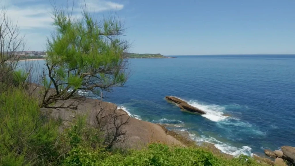 Blick von einer Klippe hinab auf das tiefblaue Meer der kantabrischen See. Wellen brechen sich weiß schäumend an einer markanten, länglichen Felsnadel im Wasser. Im linken Vordergrund wachsen grüne Büsche.